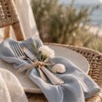 Coastal Wedding Tischdeko mit Keramikteller, taubenblauer Leinenservierte, Muschel und gebundenem Besteck auf einem Rattanstuhl am Meer.