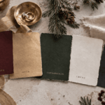 Fünf Farbkarten (Bordeaux, Gold, Tannengrün, Creme, Schwarz) umgeben von Rose, Ringschale, Tannenzweig, Leinenstoff und dunklem Teller.